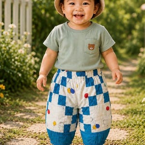 May include: A smiling baby wearing a light green t-shirt with a bear pocket detail and a beige bear-eared hat. The baby is also wearing blue and white checkered pants with balloon and bear designs.
