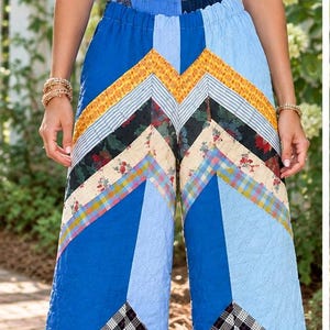 May include: A white ribbed tank top paired with wide-leg quilted pants featuring a vibrant patchwork design. The pants display a chevron pattern composed of various fabric strips in shades of blue, yellow, red, black, and white with floral and plaid prints.