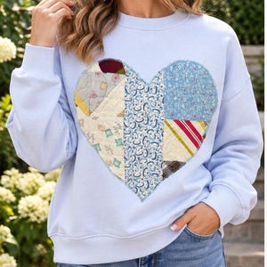 May include: A light blue crewneck sweatshirt featuring a large, central heart-shaped applique. The heart is constructed from a patchwork of various quilted fabric patterns, including floral, striped, and geometric designs in muted tones.