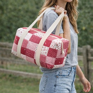 May include: A red and white patchwork duffel bag with cream-colored straps and a zipper closure. The bag features a variety of red and white patterned squares. The bag is being carried over the shoulder.