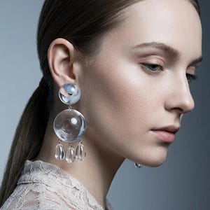 May include: Elegant, clear glass earrings with a unique design. Each earring has a small, round top with a pink and blue pattern, a large, clear sphere, and three clear, teardrop-shaped dangles. The earrings are worn by a person with dark hair.