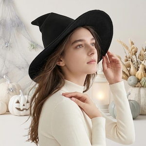 Puede incluir: Un sombrero de bruja negro con ala ancha y copa puntiaguda. La persona lleva un cuello alto blanco. El fondo incluye calabazas blancas, arañas decorativas y una linterna, sugiriendo una temática de Halloween.