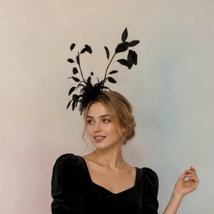 May include: A black fascinator headpiece featuring a cluster of dark feathers and thin, upright stems with leaf-like accents. The accessory is worn on the head, showcasing a structured arrangement of black plumage that extends upwards and outwards.