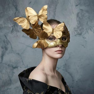 May include: A gold sequined mask with butterfly accents. The mask covers the eyes and is adorned with multiple gold butterfly wings. The woman is wearing a black dress with gold embroidery. The background is a mottled grey and blue.