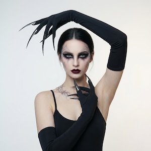 May include: A person wearing a black tank top and long black gloves with long, pointed, black fingernails. The person has dark eye makeup and dark lipstick. A tattoo is visible on the person's neck.