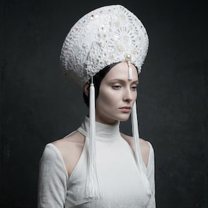 Peut inclure: Une coiffe de style kokochnik blanche ornée de dentelle complexe, de paillettes scintillantes et de petites perles. La couronne haute et arrondie présente un détail central en perle et de longs pompons blancs délicats sur les côtés.