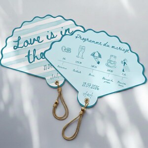 Peut inclure: Deux programmes de mariage en forme d'éventail bleu clair. L'un présente des rayures bleues et blanches et le texte "Love is in the" avec la date 21.07. L'autre présente un calendrier de mariage avec des illustrations et le texte "Programme du mariage" et la date 21.07.2026. Les deux ont des glands dorés.