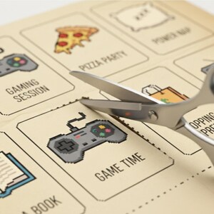 May include: A sheet of paper with pixelated illustrations of leisure activities, including a pizza slice, a controller labeled "Game Time," and a book. Scissors are cutting out the "Game Time" illustration.