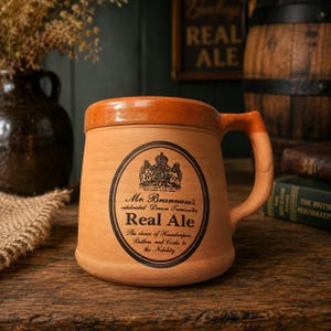 May include: A terracotta mug with a smooth orange-brown rim and handle. The body features a black oval label with a crest and text reading: Mr. Brannam's celebrated Devon Terracotta Real Ale, The choice of Housekeeper, Butlers and Cooks to the Nobility.