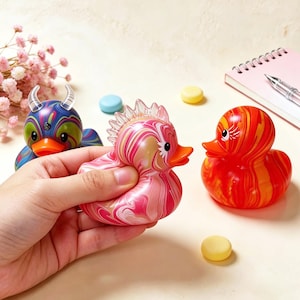 May include: A collection of decorative rubber ducks in various colors and designs. One duck is held in a hand, featuring pink and white swirls. Another is red and orange, and a third is blue with horns.