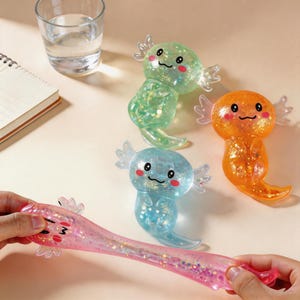 Könnte beinhalten: Vier quetschbare Spielzeuge in Axolotl-Form aus einem durchscheinenden, mit Glitzer gefüllten Material. Die Figuren sind grün, orange, blau und rosa und haben schwarze Augen, einen kleinen Mund, rosa Wangenflecken und äußere Kiemen.