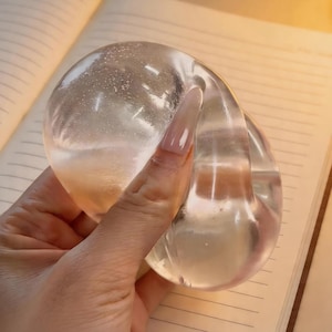 May include: A clear, amorphous glass sculpture with a smooth, reflective surface. The sculpture is held in a hand, and the light catches its curves, creating highlights and shadows. The background is a notebook with lined pages.