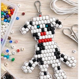 May include: A handmade dog keychain made from white, black, and red pony beads. The keychain is attached to a silver clasp. The image also shows containers of colorful pony beads, scissors, and the text "DOG KEYCHAIN PATTERNS Pony Bead PDF".