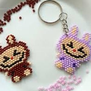 May include: Two beaded bunny keyrings, one brown and one purple, with black and tan details. The keyrings are on a white surface, surrounded by scattered beads. Each bunny has a smiling face.