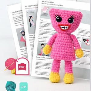 Op de afbeelding: Een handgemaakte roze gehaakte pop met zwarte ogen, wimpers en een rode lachende mond. De pop heeft gele handen en voeten. Verschillende vellen papier met illustraties en tekst op de achtergrond.