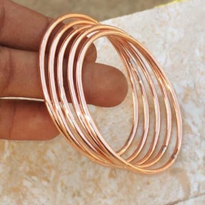 以下が含まれることがあります： 銅色のバングルセット。バングルは丸く、滑らかで磨かれた仕上がりです。ジュエリーは明るい色の表面に表示されています。バングルは暖かくメタリックな色です。