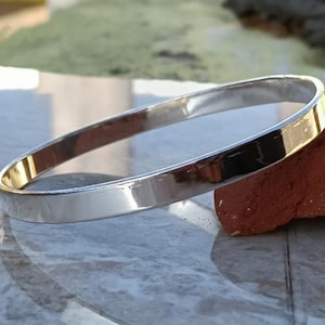 Puede incluir: Un brazalete plateado brillante, de forma rectangular. El brazalete está sobre una superficie reflectante, junto a un pequeño ladrillo rojizo. Esta joya está destinada a ser usada en la muñeca.