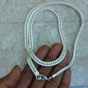 May include: A silver-colored chain necklace with a woven design. The necklace is made of a shiny, metallic material and features a lobster clasp closure. The chain is displayed on a neutral background.