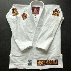 Puede incluir: Kimono de Jiu-Jitsu blanco con logotipos bordados en morado y dorado. El kimono presenta la marca "Shoyoroll" en el pecho, los hombros y la parte inferior delantera. El kimono está hecho de tela acanalada.