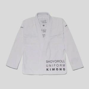May include: A white martial arts uniform, or gi, with the words "SHOYOROLL UNIFORM KIMONO" printed in black on the front. The gi has long sleeves and a collar, and is laid flat against a light grey background.