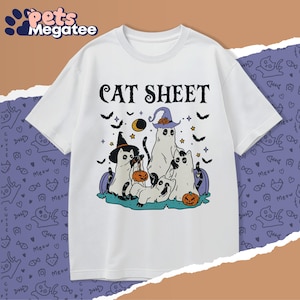 May include: White t-shirt with a Halloween-themed graphic. The design features cartoon cats dressed as ghosts, wearing witch hats, and holding pumpkins. The text "CAT SHEET" is printed above the illustration, with bats and stars scattered around.