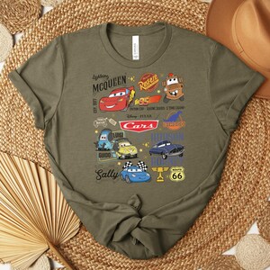Puede incluir: Camiseta verde oliva con un gr&aacute;fico colorido de personajes y logotipos de la pel&iacute;cula Cars de Disney Pixar. El dise&ntilde;o incluye a Rayo McQueen, Mate y otros personajes, junto con el letrero de la Ruta 66.