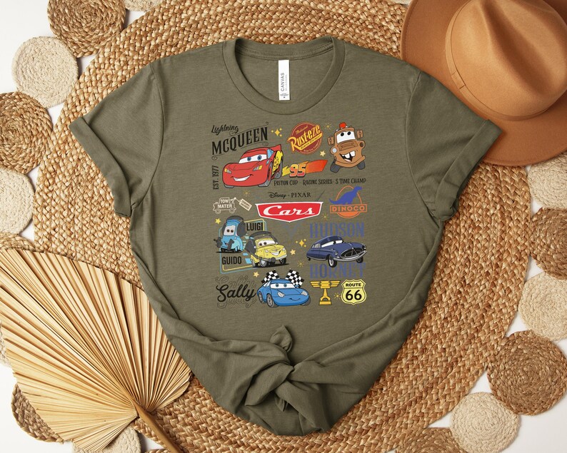Puede incluir: Camiseta verde oliva con un gr&aacute;fico colorido de personajes y logotipos de la pel&iacute;cula Cars de Disney Pixar. El dise&ntilde;o incluye a Rayo McQueen, Mate y otros personajes, junto con el letrero de la Ruta 66.
