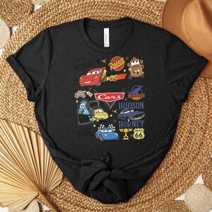 Puede incluir: Camiseta negra con un gr&aacute;fico colorido que muestra personajes y logotipos de la pel&iacute;cula "Cars". El dise&ntilde;o incluye a Rayo McQueen, Mate y otros personajes, junto con el logotipo de "Cars" y la se&ntilde;alizaci&oacute;n de la Ruta 66.