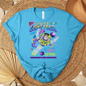 May include: A turquoise blue t-shirt featuring a graphic of Buzz Lightyear in a space suit. The design includes the words "Buzz Lightyear" in a neon font, along with the phrases "Space Ranger" and "Prepare for Ignition."