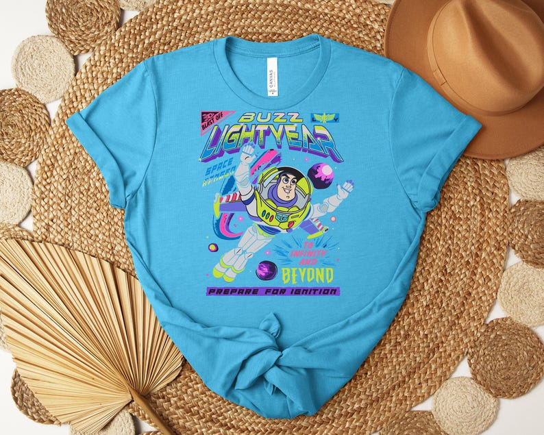 May include: A turquoise blue t-shirt featuring a graphic of Buzz Lightyear in a space suit. The design includes the words "Buzz Lightyear" in a neon font, along with the phrases "Space Ranger" and "Prepare for Ignition."
