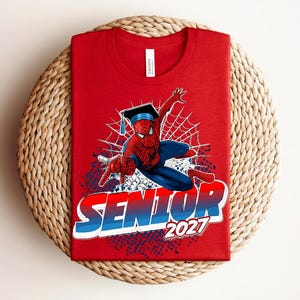 May include: A red crew neck t-shirt featuring a graphic of a superhero wearing a black graduation cap. The design includes a white spider web background and the bold text SENIOR 2027 in a red, white, and blue gradient color scheme.