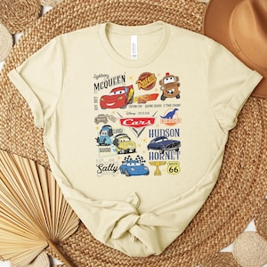 Puede incluir: Una camiseta de color beige claro con un gr&aacute;fico colorido de personajes y logotipos de la pel&iacute;cula de Disney Pixar "Cars". El dise&ntilde;o incluye a Rayo McQueen, Mate y otros personajes, junto con el texto "Cars" y otros textos relacionados.