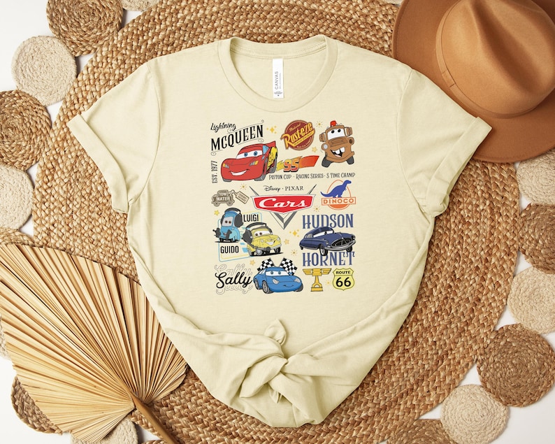 Puede incluir: Una camiseta de color beige claro con un gr&aacute;fico colorido de personajes y logotipos de la pel&iacute;cula de Disney Pixar "Cars". El dise&ntilde;o incluye a Rayo McQueen, Mate y otros personajes, junto con el texto "Cars" y otros textos relacionados.