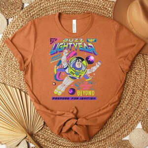 May include: An orange t-shirt featuring a graphic of Buzz Lightyear in a space suit. The design includes the text "Buzz Lightyear" in a colorful, retro font, along with the phrases "Blast Off" and "Beyond". The shirt is made of a soft material.