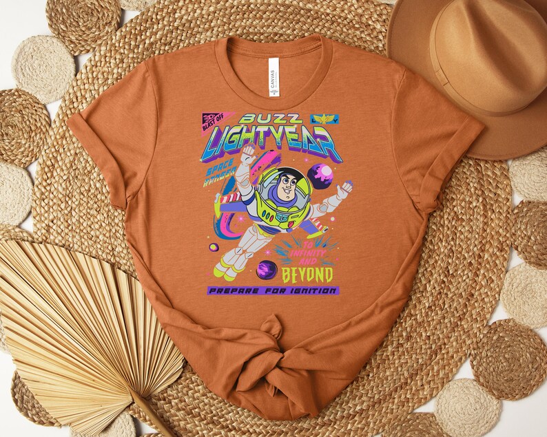 May include: An orange t-shirt featuring a graphic of Buzz Lightyear in a space suit. The design includes the text "Buzz Lightyear" in a colorful, retro font, along with the phrases "Blast Off" and "Beyond". The shirt is made of a soft material.
