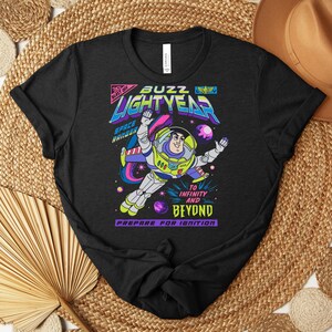 May include: Black t-shirt featuring a colorful graphic of Buzz Lightyear in a space scene. The design includes the words "Buzz Lightyear", "Space Ranger", and "To Infinity and Beyond". The shirt is a crew neck.