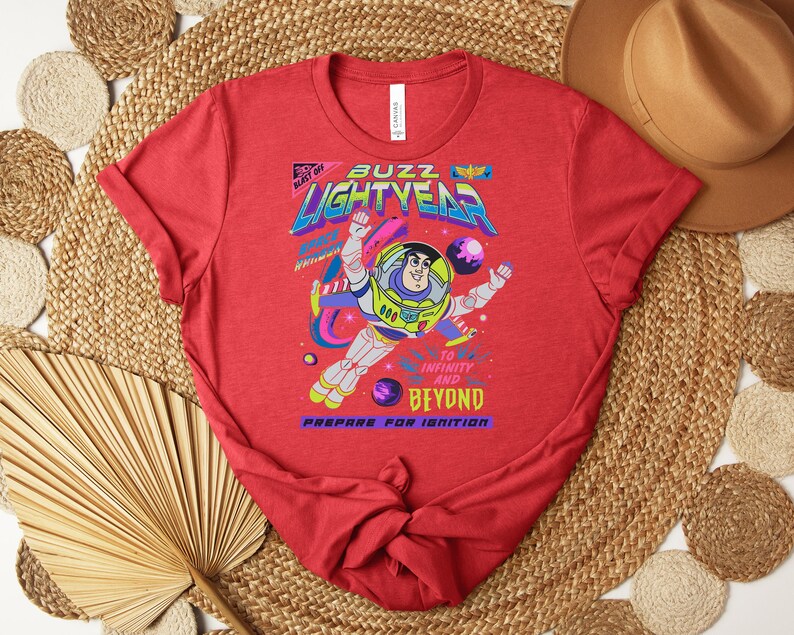 May include: Red t-shirt featuring a graphic of Buzz Lightyear in a spacesuit, with the words "Buzz Lightyear" and "To Infinity and Beyond." The design includes planets and stars, with the phrase "Prepare for Ignition."