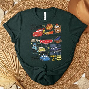 Puede incluir: Camiseta verde oscuro con un gr&aacute;fico colorido que muestra personajes y logotipos de la pel&iacute;cula animada "Cars". El dise&ntilde;o incluye a Rayo McQueen, Mate y otros personajes, junto con el logotipo de "Cars" y la se&ntilde;alizaci&oacute;n de la Ruta 66.