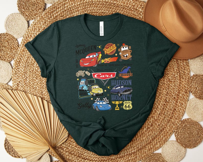 Puede incluir: Camiseta verde oscuro con un gr&aacute;fico colorido que muestra personajes y logotipos de la pel&iacute;cula animada "Cars". El dise&ntilde;o incluye a Rayo McQueen, Mate y otros personajes, junto con el logotipo de "Cars" y la se&ntilde;alizaci&oacute;n de la Ruta 66.