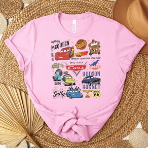 Puede incluir: Una camiseta rosa claro con un dise&ntilde;o gr&aacute;fico colorido con personajes y logotipos de la pel&iacute;cula animada "Cars". El dise&ntilde;o incluye a Rayo McQueen, Sally y otros personajes, junto con el texto "Cars" y "Ruta 66".