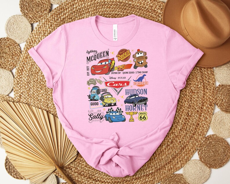 Puede incluir: Una camiseta rosa claro con un dise&ntilde;o gr&aacute;fico colorido con personajes y logotipos de la pel&iacute;cula animada "Cars". El dise&ntilde;o incluye a Rayo McQueen, Sally y otros personajes, junto con el texto "Cars" y "Ruta 66".