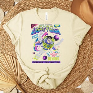 May include: A light yellow t-shirt featuring a colorful graphic of Buzz Lightyear in a dynamic pose. The design includes the text "Buzz Lightyear" and "To Infinity and Beyond". The shirt is a classic crew neck style.