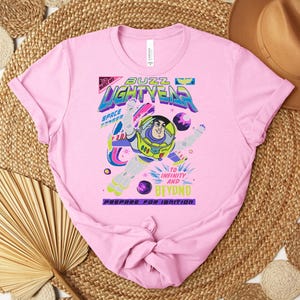 May include: A light pink t-shirt featuring a graphic of Buzz Lightyear with the words "Buzz Lightyear" in a colorful, futuristic font. The design includes the phrases "To Infinity and Beyond" and "Prepare for Ignition."