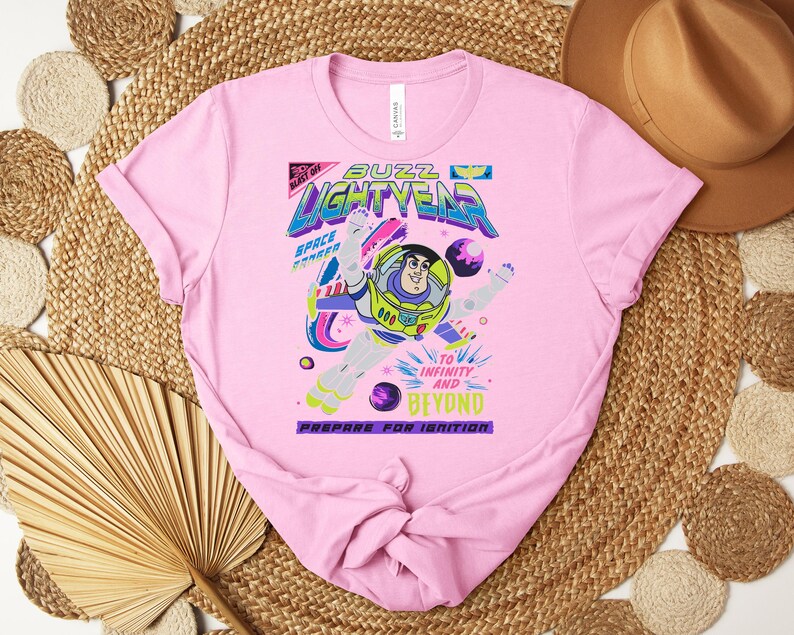 May include: A light pink t-shirt featuring a graphic of Buzz Lightyear with the words "Buzz Lightyear" in a colorful, futuristic font. The design includes the phrases "To Infinity and Beyond" and "Prepare for Ignition."
