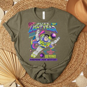 May include: Olive green t-shirt featuring a graphic of Buzz Lightyear in a space suit, with the words "Buzz Lightyear" and "To Infinity and Beyond." The design includes planets and a colorful space theme.