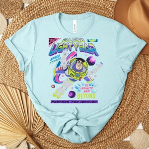 May include: Light blue t-shirt featuring a graphic of Buzz Lightyear from Toy Story. The design includes the text "Buzz Lightyear" and "To Infinity and Beyond." The shirt has a crew neck and short sleeves.