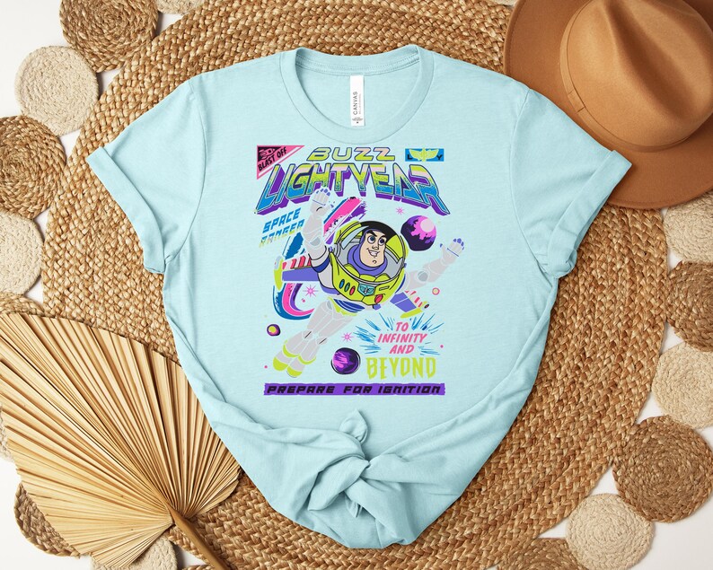 May include: Light blue t-shirt featuring a graphic of Buzz Lightyear from Toy Story. The design includes the text "Buzz Lightyear" and "To Infinity and Beyond." The shirt has a crew neck and short sleeves.