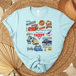 Puede incluir: Camiseta azul claro con un gr&aacute;fico colorido de personajes y logotipos de la pel&iacute;cula animada "Cars". El dise&ntilde;o incluye a Rayo McQueen, Mate y otros personajes, junto con el logotipo de "Cars" y la se&ntilde;al de la Ruta 66.