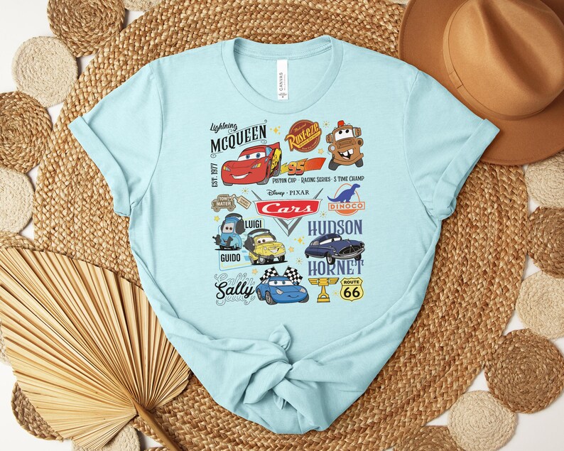 Puede incluir: Camiseta azul claro con un gr&aacute;fico colorido de personajes y logotipos de la pel&iacute;cula animada "Cars". El dise&ntilde;o incluye a Rayo McQueen, Mate y otros personajes, junto con el logotipo de "Cars" y la se&ntilde;al de la Ruta 66.