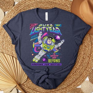 May include: Navy blue t-shirt featuring a colorful graphic of Buzz Lightyear in a space scene. The design includes the text "Buzz Lightyear" and "To Infinity and Beyond." The shirt has a crew neck and short sleeves.